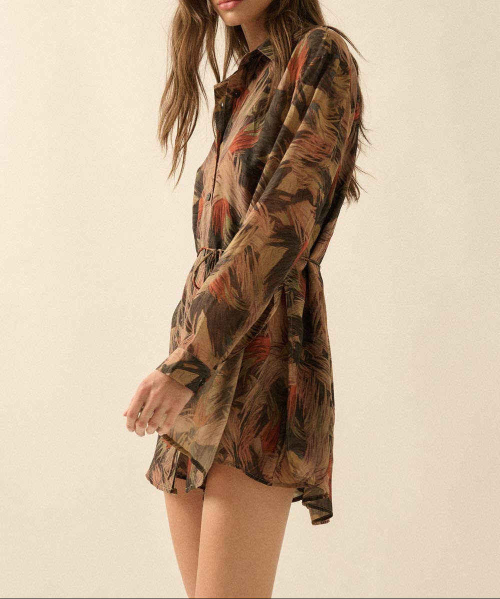A woman modeling a long-sleeve button-up shirt dress with a brown, beige, and rust-toned abstract print against a plain beige studio background.