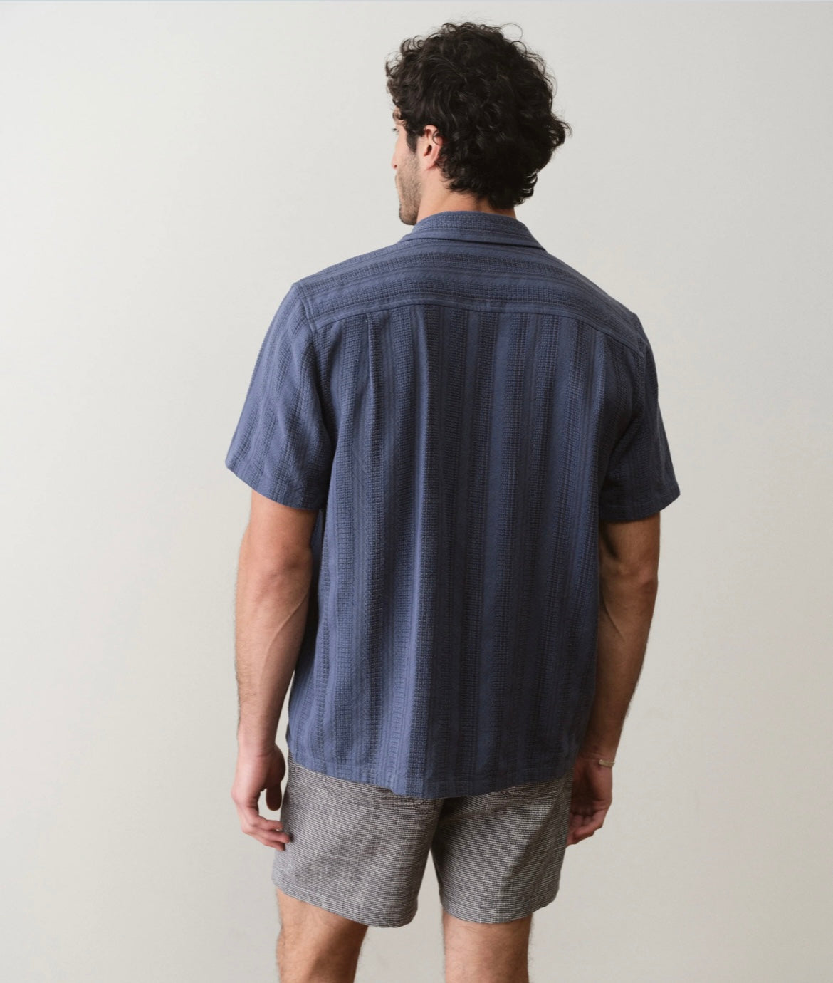 Man wearing a blue short-sleeve shirt and gray shorts on a plain background