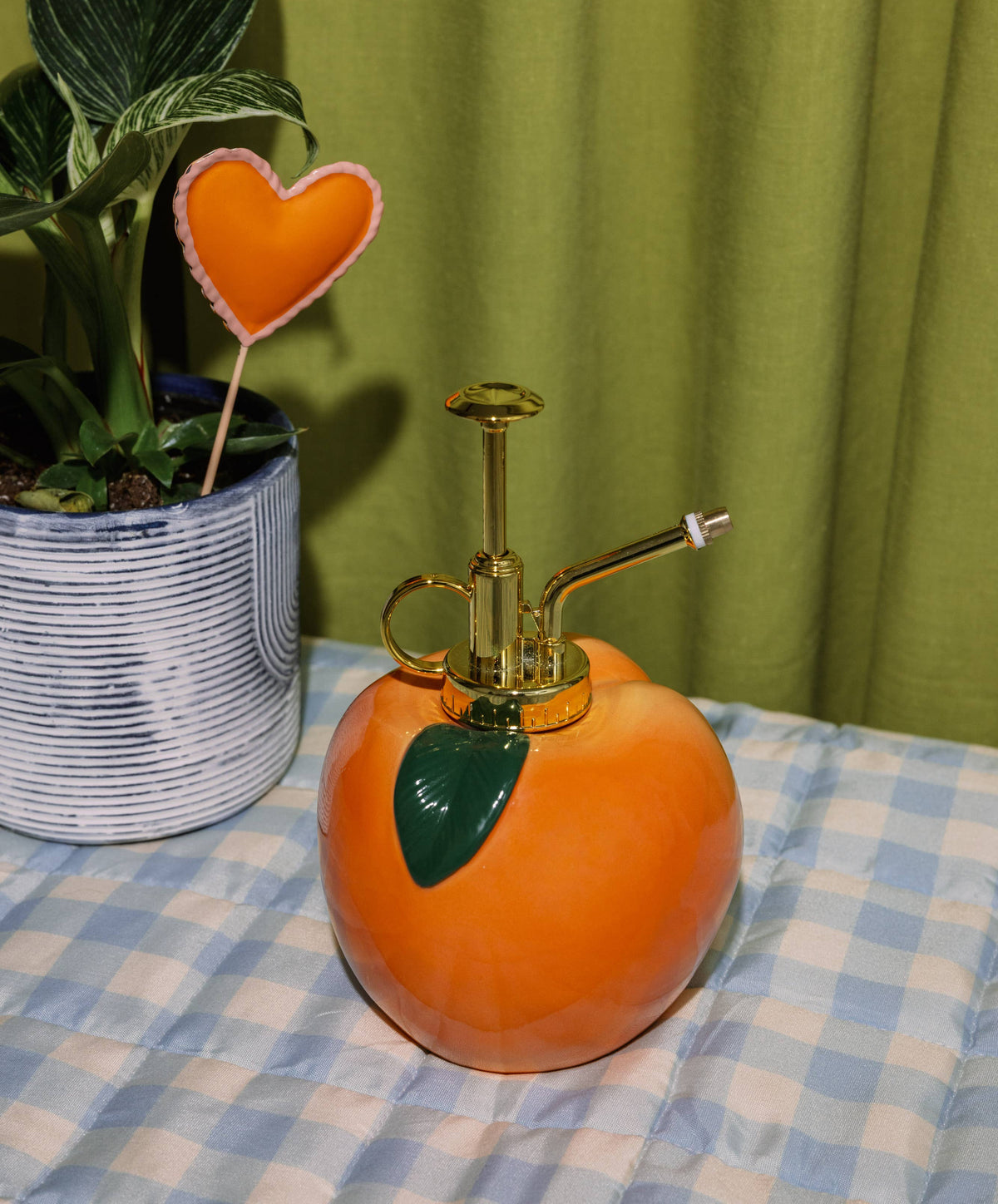 Orange spray bottle with gold nozzle on a checkered tablecloth, next to a potted plant against a green curtain.