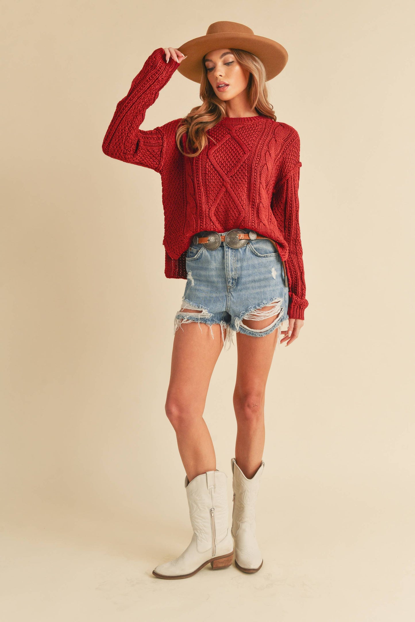 Woman wearing a red sweater, denim shorts, and white cowboy boots on a beige background