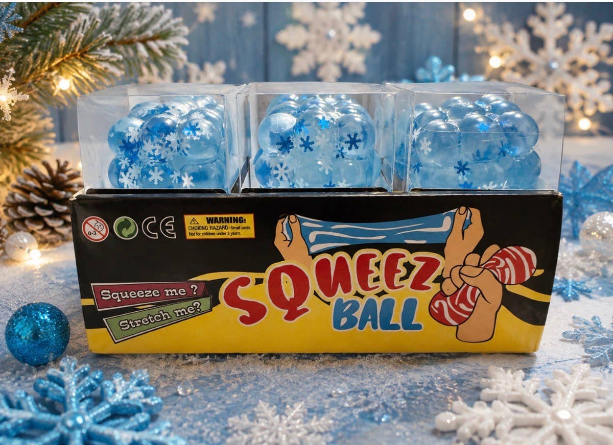 A boxed toy set labeled 'SQUEEZ BALL' with three blue translucent squishy balls inside clear plastic packaging, displayed on a festive snowy surface with pine branches and lights in the background.