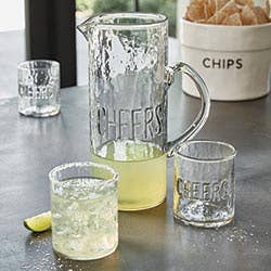 Clear 'CHEERS' glass pitcher and tumblers on a table with chips in the background
