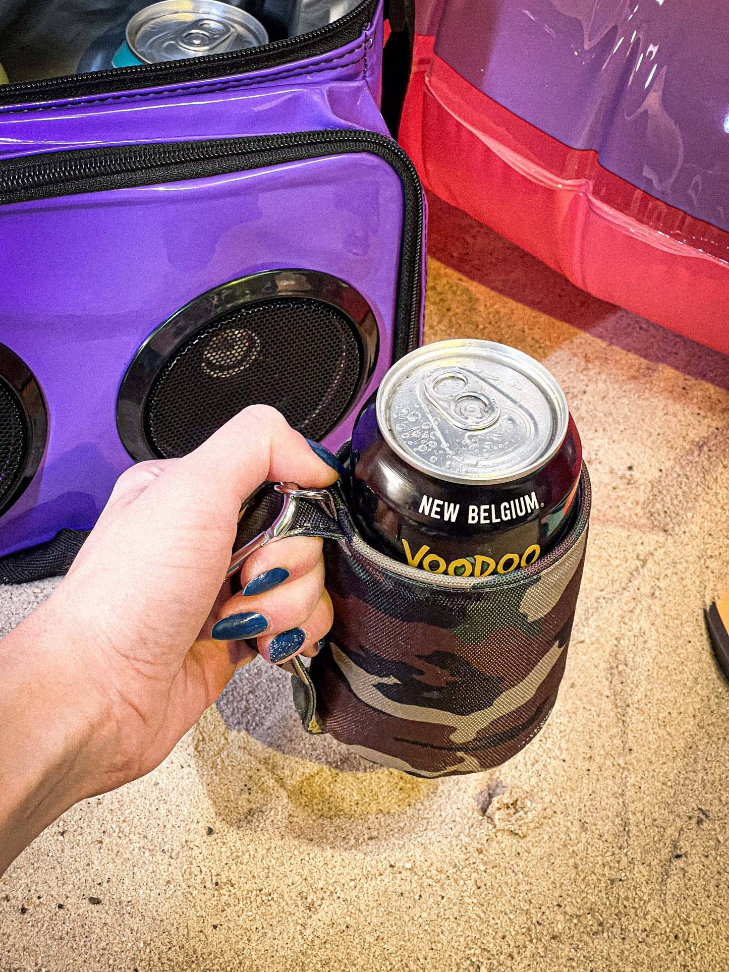 Person holding a can of New Belgium Voodoo beer with a camo holder next to a purple cooler.
