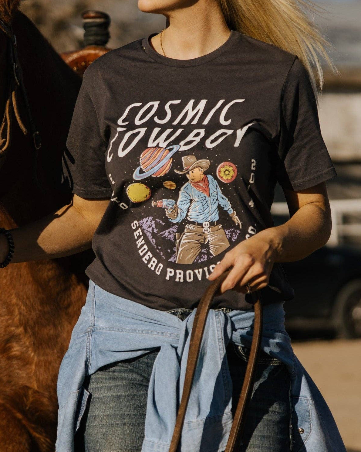 Woman wearing a 'Cosmic Cowboy' t-shirt standing next to a horse in an outdoor setting.