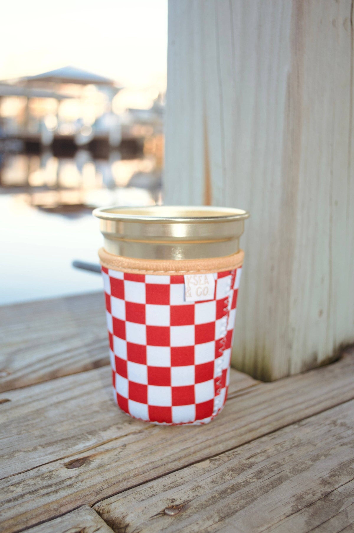 A red and white checkered reusable cup with a gold metal lid and a tan fabric sleeve, sitting on a weathered wooden deck with a blurred outdoor background.