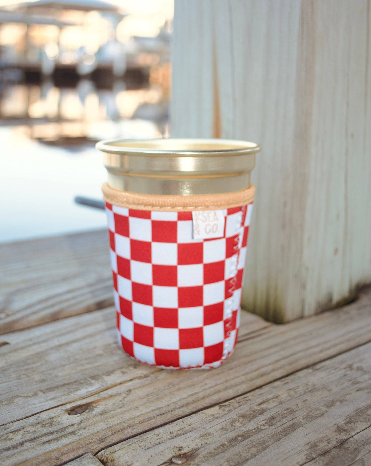 A red and white checkered reusable cup with a gold metal lid and a tan fabric sleeve, sitting on a weathered wooden deck with a blurred outdoor background.
