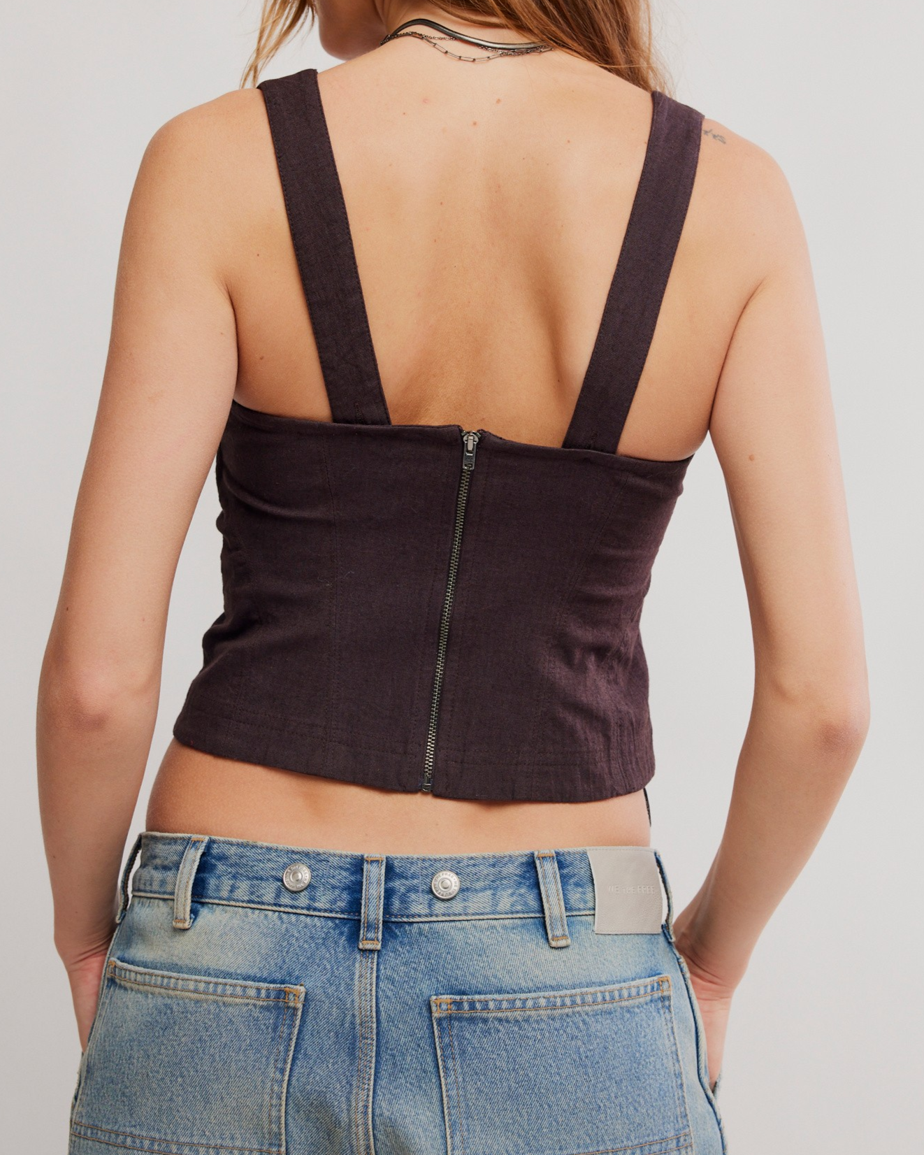 Rear view of a woman wearing a dark plum purple ribbed knit crop top with wide shoulder straps and a central zipper, paired with light blue denim jeans.