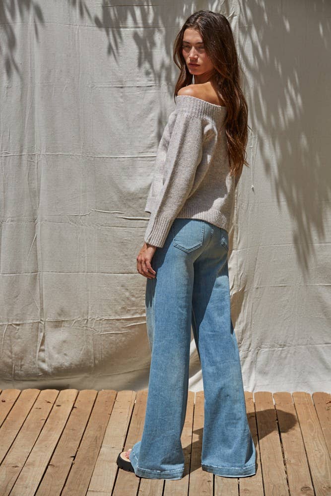 Woman wearing a light gray off-shoulder sweater and blue jeans standing against a neutral background.