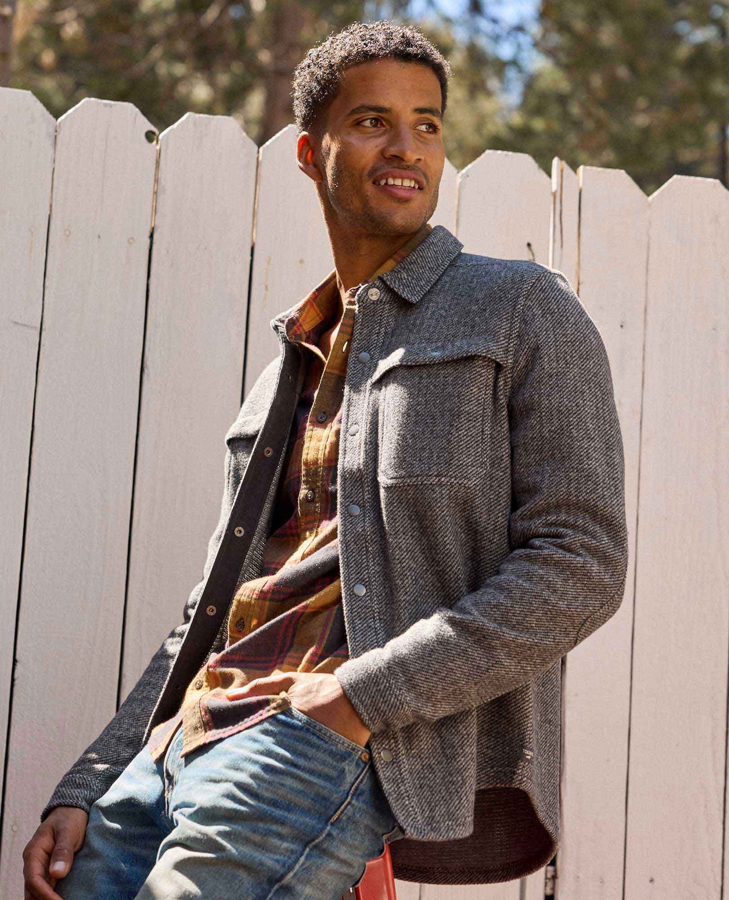 Man wearing a gray jacket and jeans standing in front of a white picket fence.
