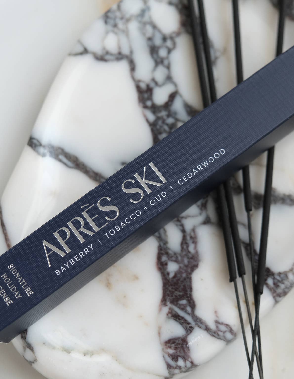 A close-up of a dark navy Apès Ski incense box placed diagonally on a white and gray marbled surface with thin black incense sticks.
