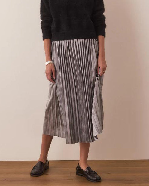 A woman standing wearing a black sweater and a midi pleated skirt with horizontal stripes. She is also wearing a watch and flat shoes.