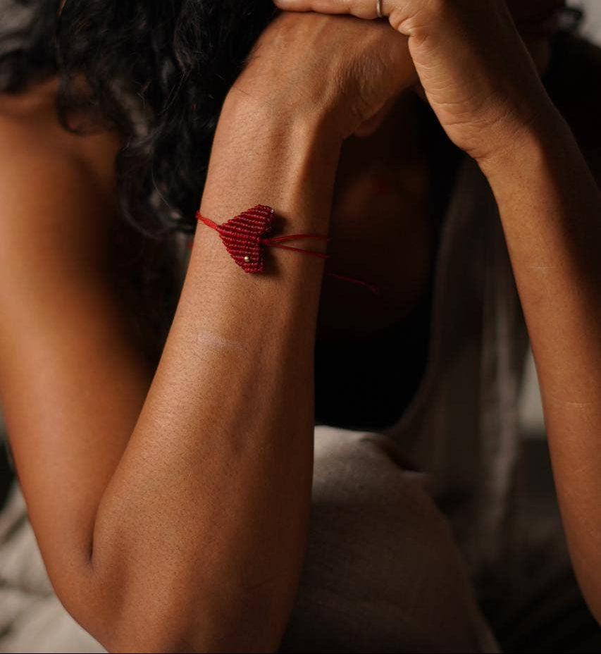 A close-up lifestyle shot of a woman with dark curly hair resting her head on her hands, showcasing a small red macrame heart bracelet on her wrist. She is wearing a purple gemstone ring on her finger.