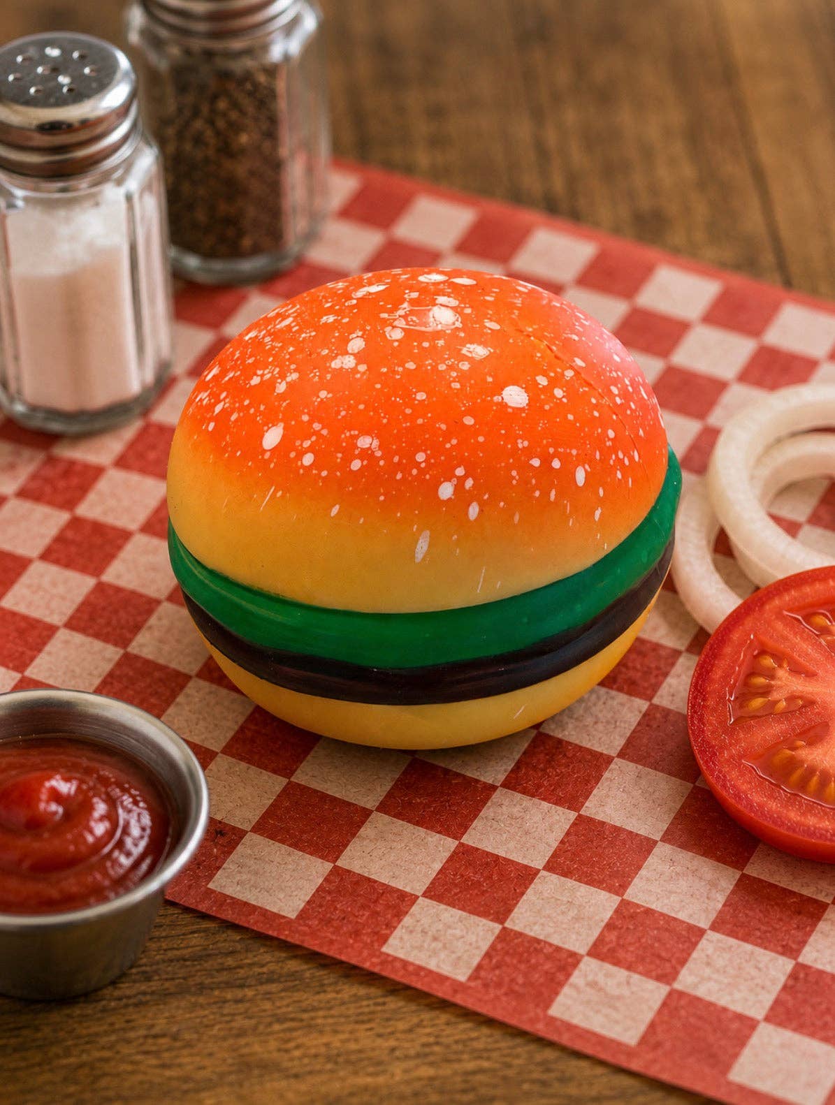 A novelty condiment container shaped like a hamburger sits on a red-and-white checkered paper mat on a wooden table, surrounded by small props including ketchup, onion rings, a tomato slice, and salt and pepper shakers.