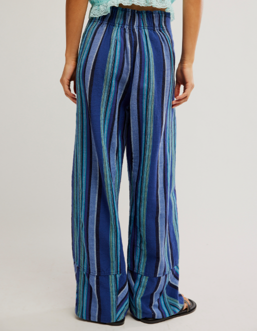 A person shown from the back wearing wide-leg pants with vertical blue, teal, and navy stripes, paired with a light green lace-trim top and black sandals, against a plain light gray studio background.