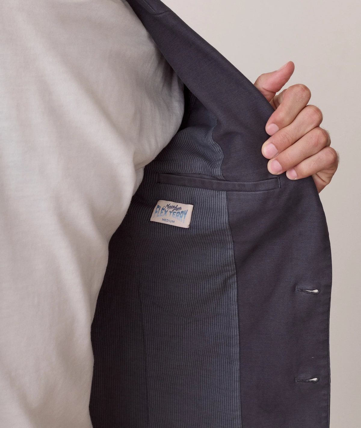 Close-up of a person wearing a dark charcoal gray pinstripe blazer over a light beige shirt, holding the lapel open to show the interior lining and a small brand label.
