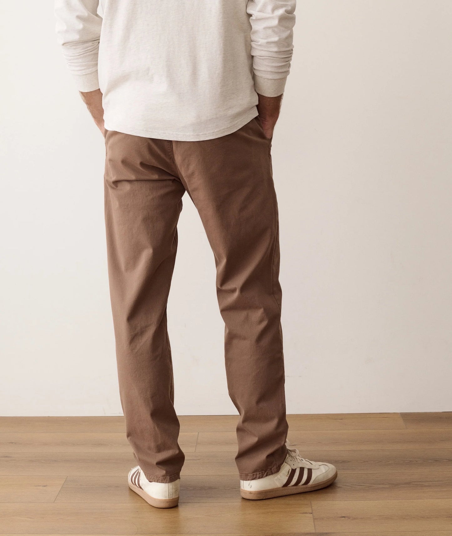 A person shown from the back wearing brown straight-leg chino pants, a white long-sleeve shirt, and white sneakers with black stripes, standing on a light wood floor against a plain off-white wall.
