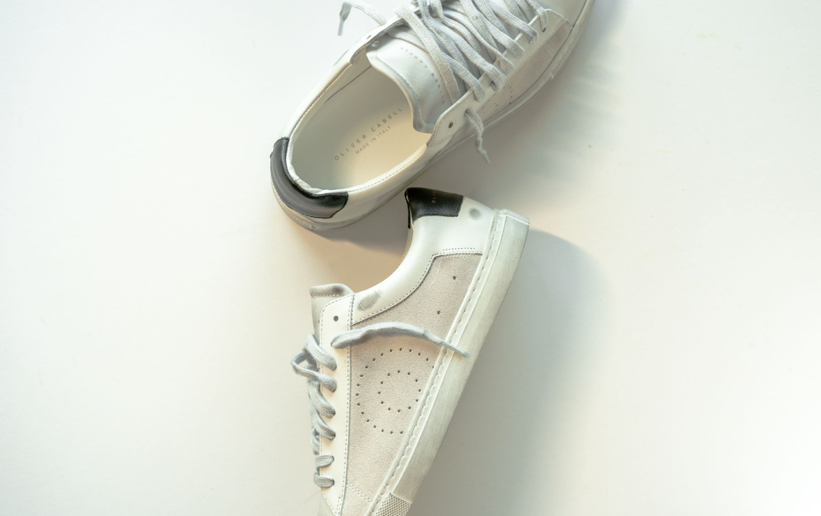 A pair of low-top sneakers in off-white and light gray, photographed from above on a plain white surface. The shoes have perforated details on the toe box and side, with white laces and a black heel accent.