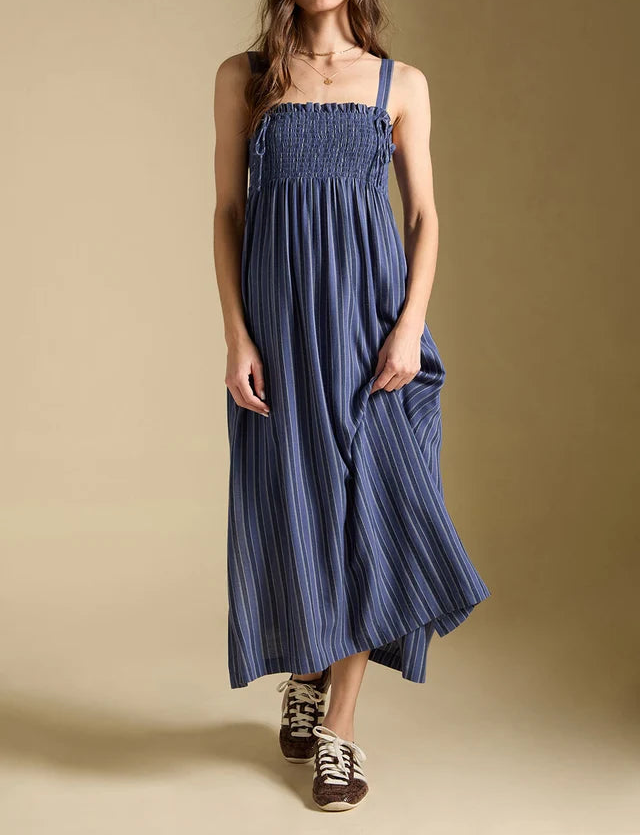 A woman modeling a blue smocked midi dress with vertical pleats, styled with a matching blue cap, gold necklace, and brown sneakers against a solid beige studio background.