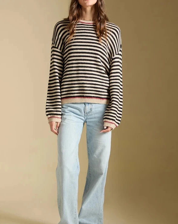 A woman modeling a black and white horizontally striped knit sweater, paired with light-wash wide-leg jeans and brown sneakers, against a solid beige studio background.