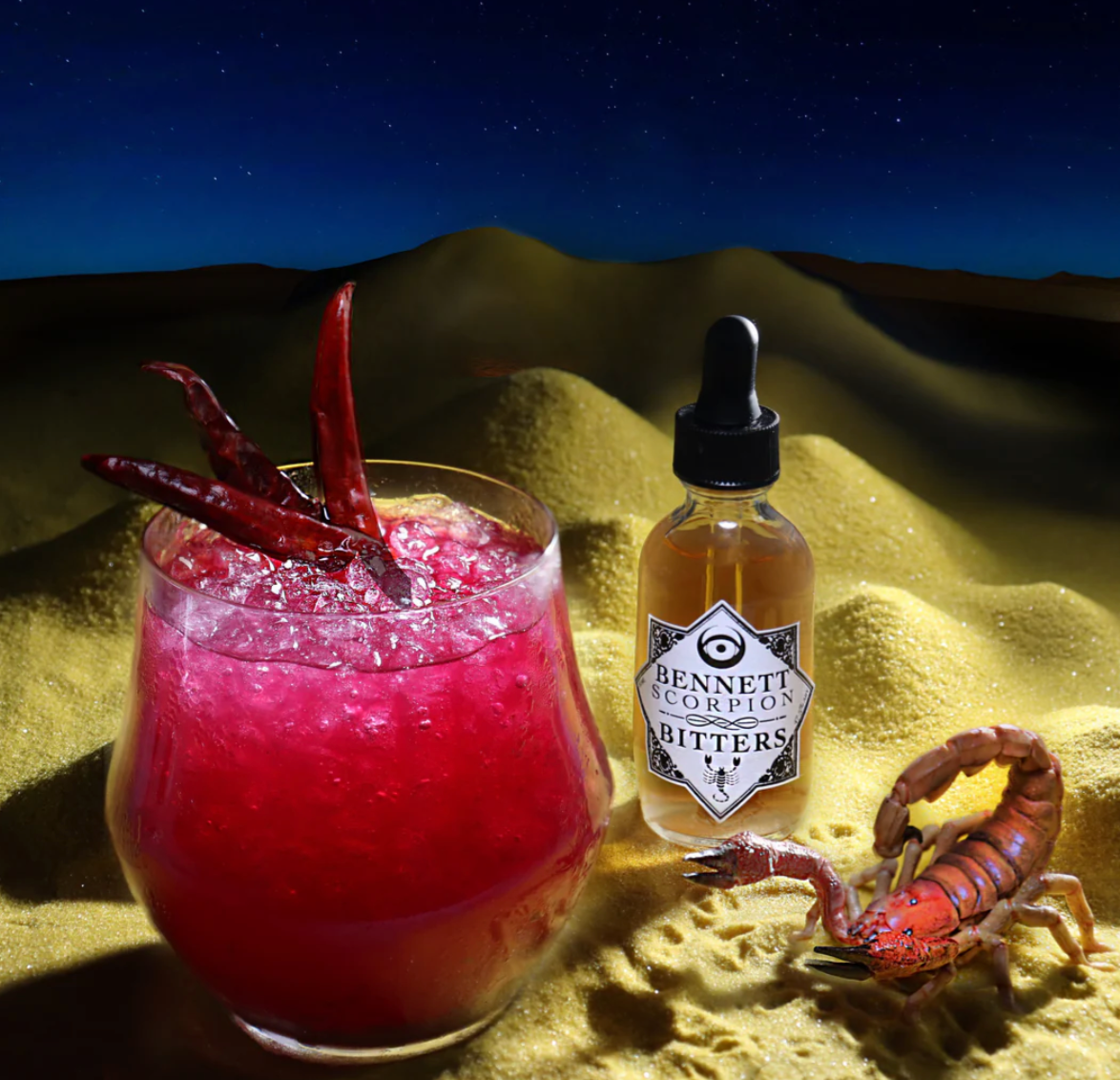 Pink cocktail with a scorpion and bottle of Bennett Scorpion Bitters on sand with a night sky.