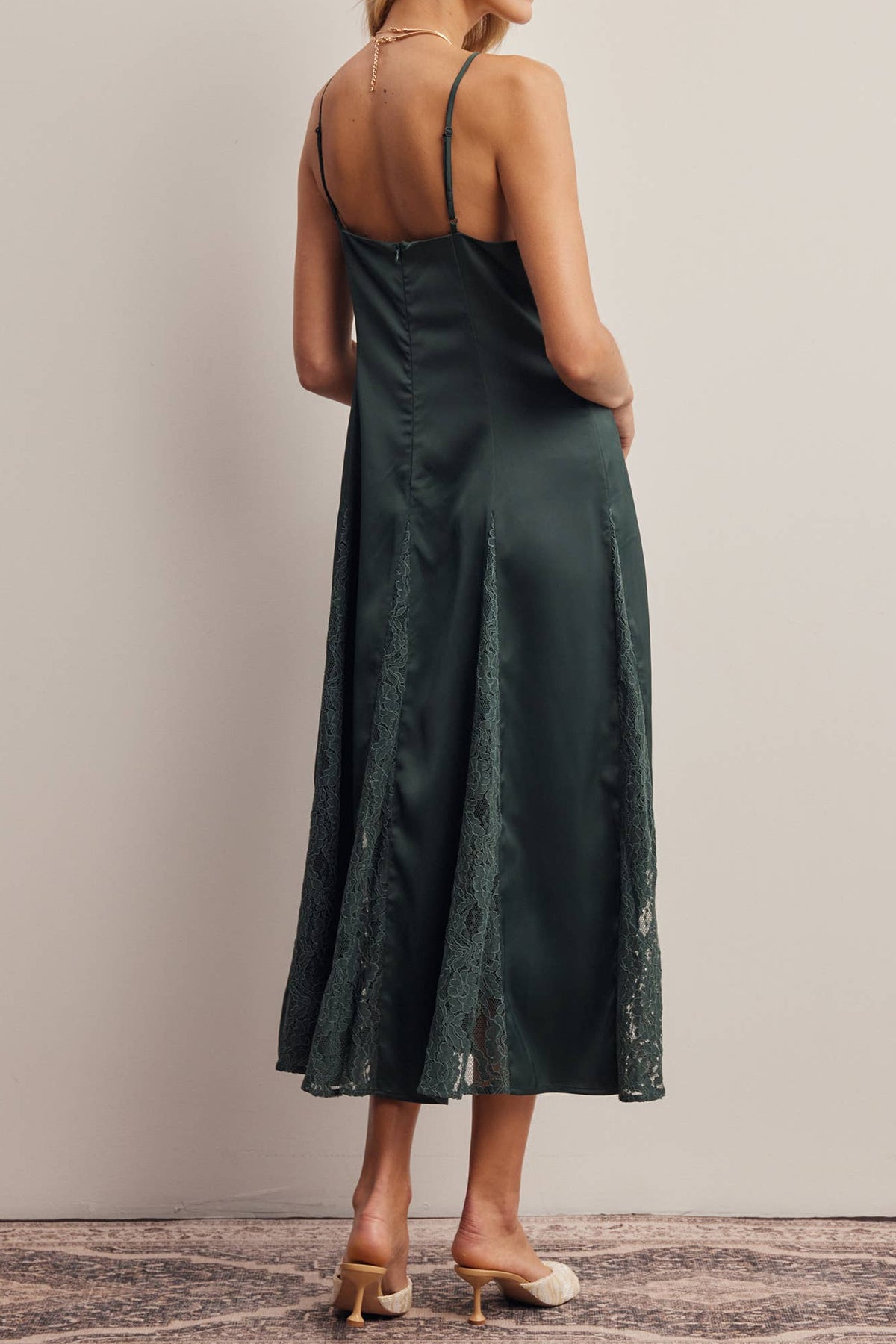 A woman modeling a dark green satin midi slip dress with lace trim, shown from the back against a neutral beige studio background.