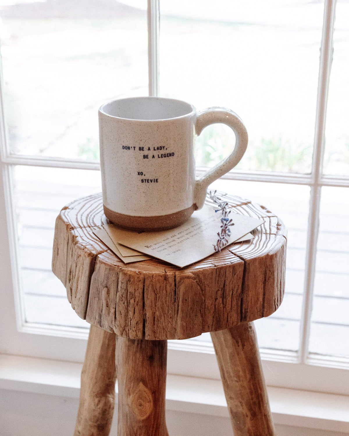A speckled cream ceramic mug with a brown base sits on a rustic wooden stool in front of a bright window. The mug has the text 'DON'T BE A LADY, BE A LEGEND XO, STEVIE' printed in black.