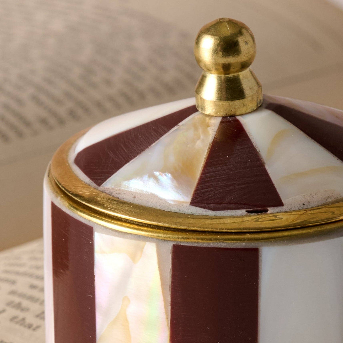 Close-up of a decorative cylindrical container with a domed lid, featuring alternating dark burgundy and white iridescent inlay panels, a gold metal rim, and a gold finial on top.