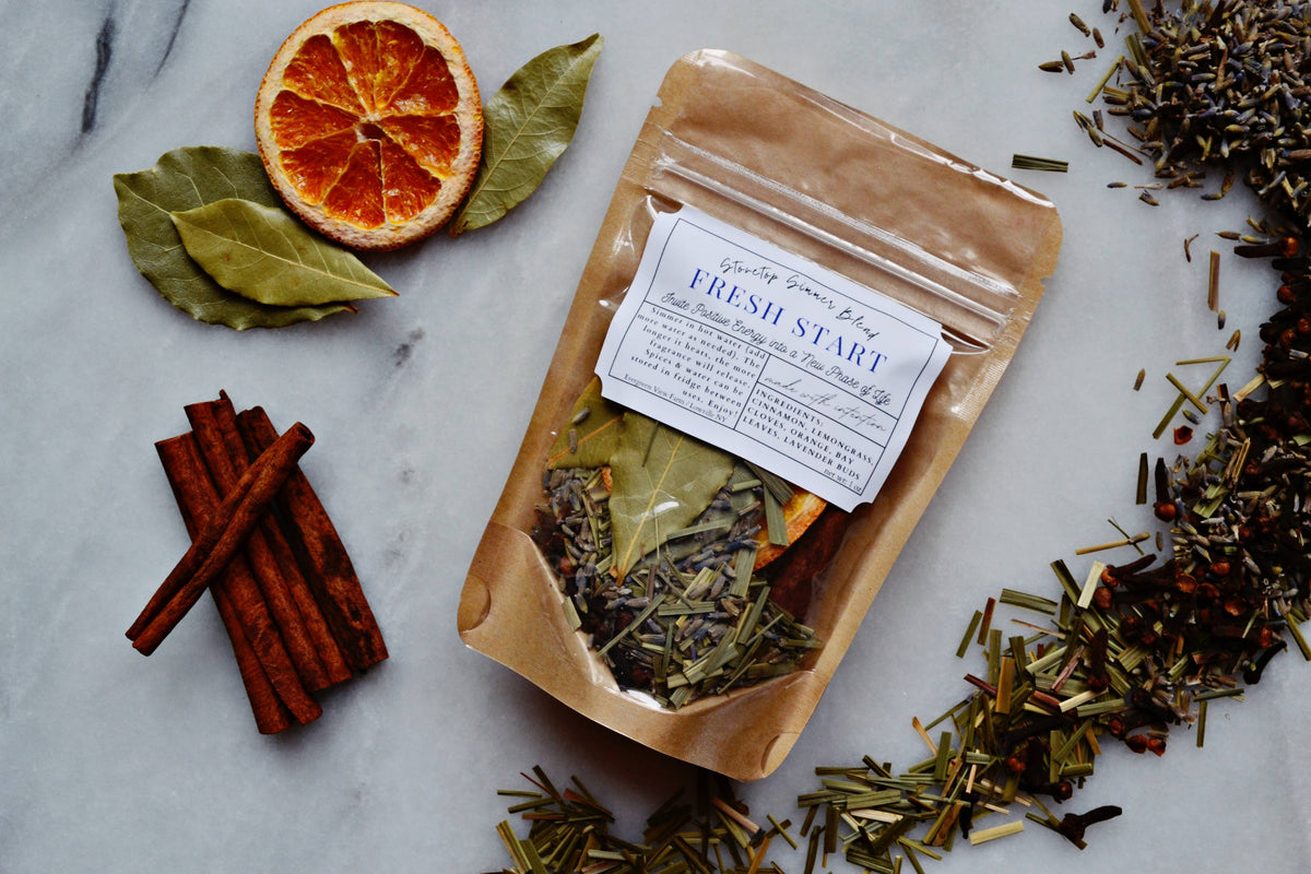 A kraft paper pouch labeled 'Fresh Start' herbal tea blend sits on a light marble surface, surrounded by loose ingredients including cinnamon sticks, bay leaves, a dried orange slice, rosemary, and other dried herbs.
