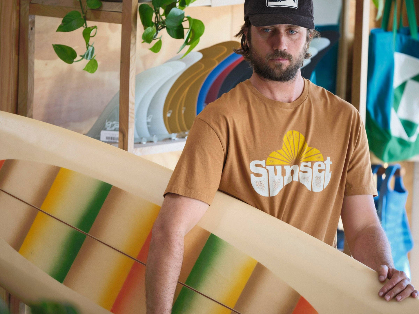 Man holding a colorful surfboard with 'Sunset' t-shirt in a store setting