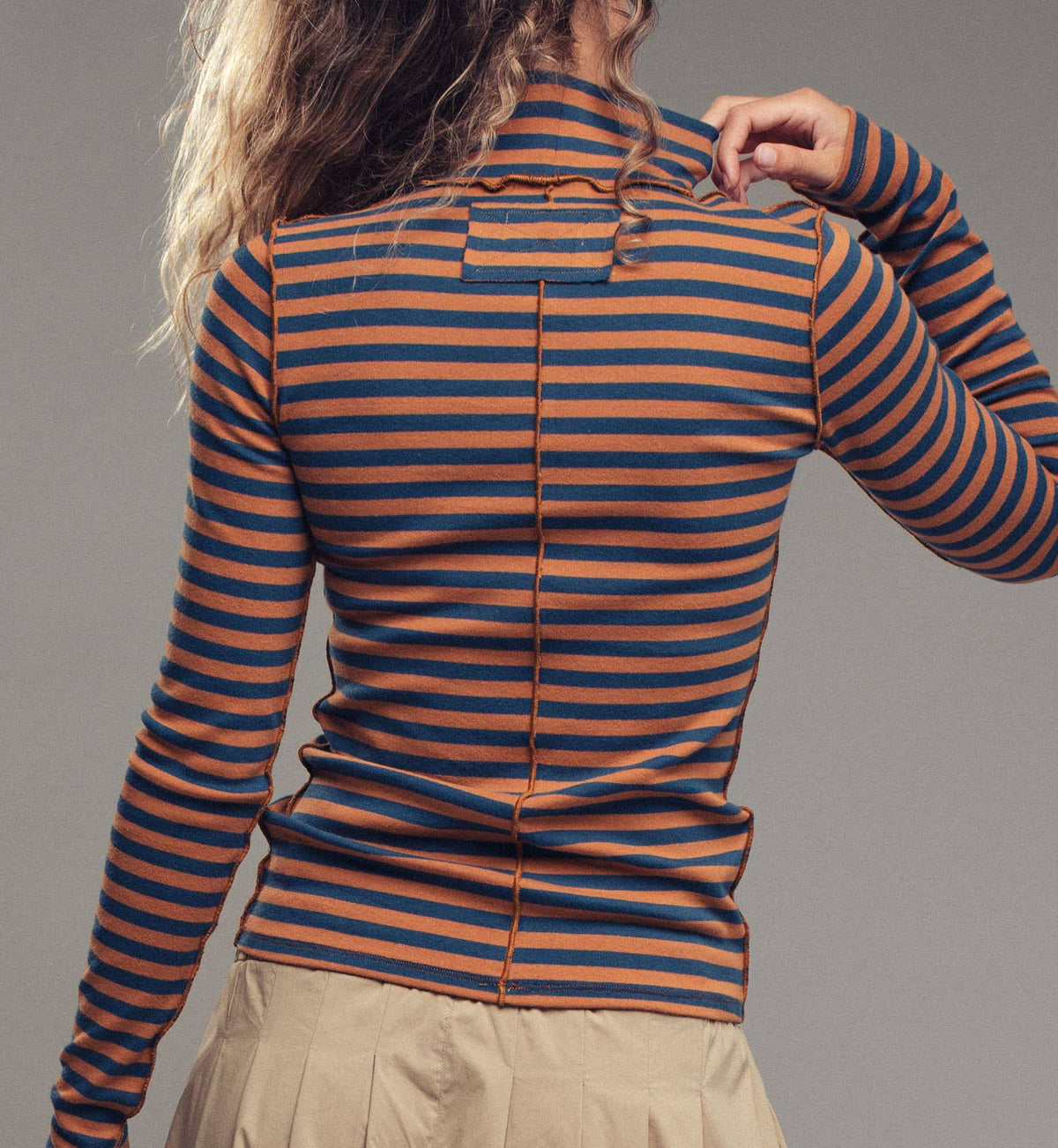 A woman shown from the back wearing a fitted long-sleeve top with horizontal rust-orange and navy stripes, paired with a beige skirt, against a plain gray studio background.