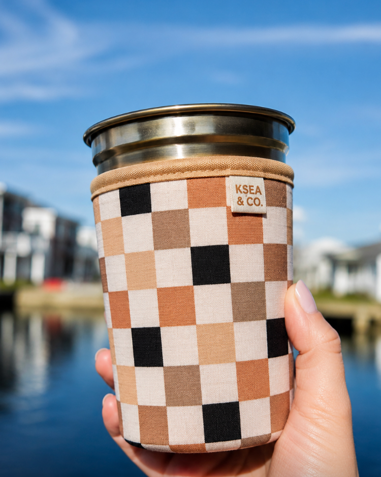 A hand holding a stainless steel tumbler with a beige and black checkerboard patterned fabric sleeve. The sleeve features a small tan leather patch with the text 'KSEA & CO.