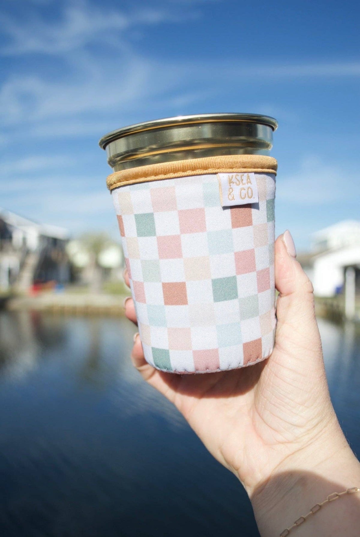 A hand holding a reusable glass can cooler with a pastel checkerboard pattern, a tan leather-like sleeve, and a clear lid, set against a blurred outdoor background of water and buildings.