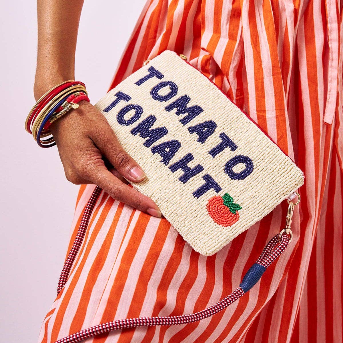 Hand holding a woven pouch with 'Tomato Tomaht' text and tomato emoji, person wearing a red and orange striped dress.