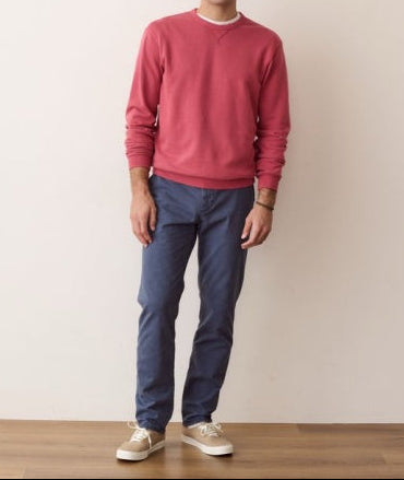 A man wearing a dusty rose crewneck sweatshirt, navy blue pants, and beige sneakers, standing against a plain off-white wall.