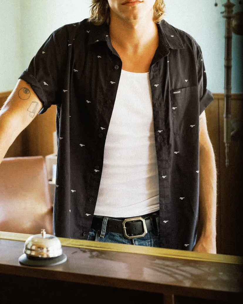 A man with long light brown hair stands indoors behind a wooden counter, wearing a black short-sleeve button-up shirt with small white embroidered details over a white tank top and blue jeans.