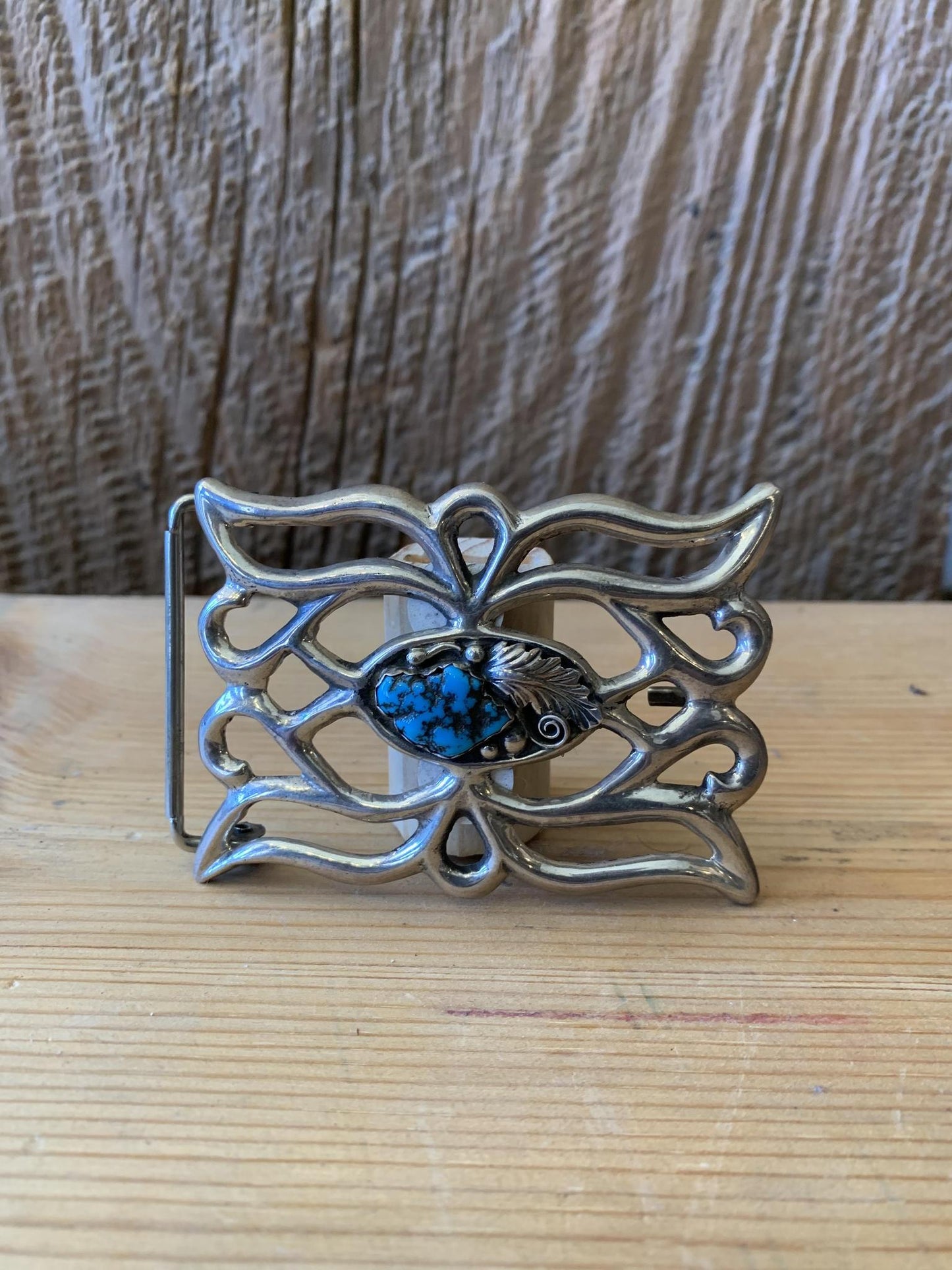 A vintage sterling silver belt buckle with turquoise detailing and a central blue stone, set against a wooden background.