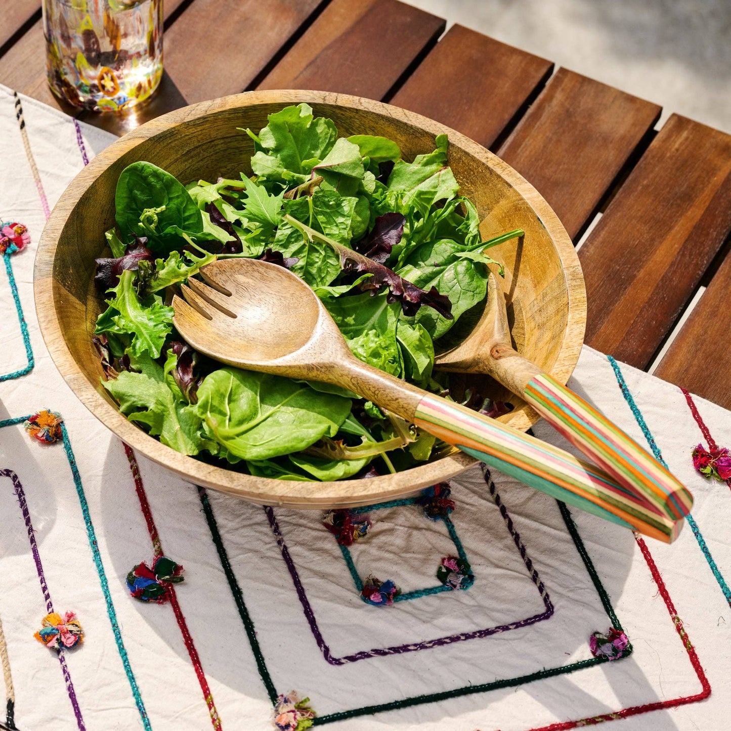 Wooden salad bowl with fresh greens and a wooden spoon on a colorful placemat.
