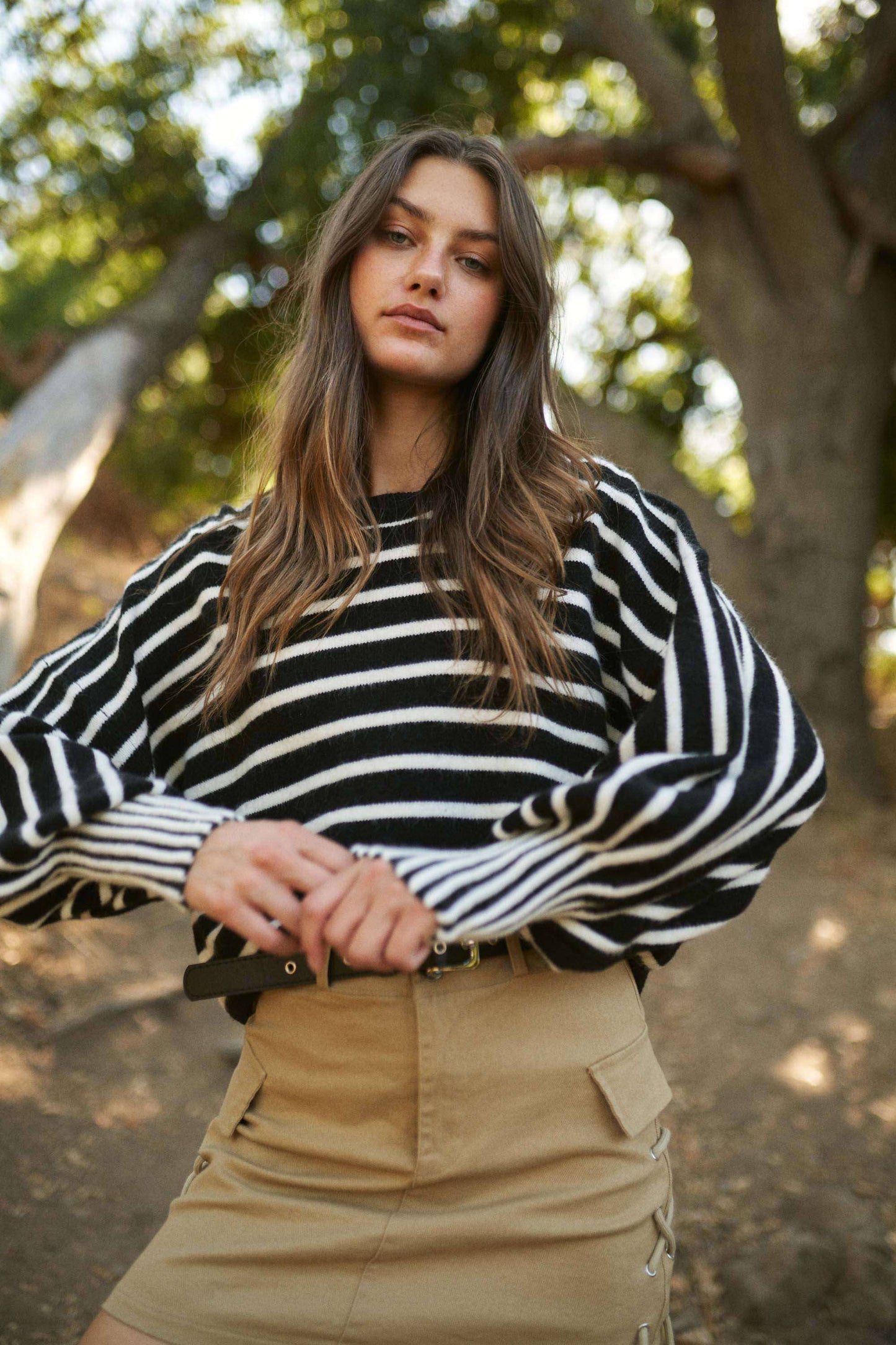 Woman wearing a black and white striped sweater and beige skirt standing outdoors with trees in the background