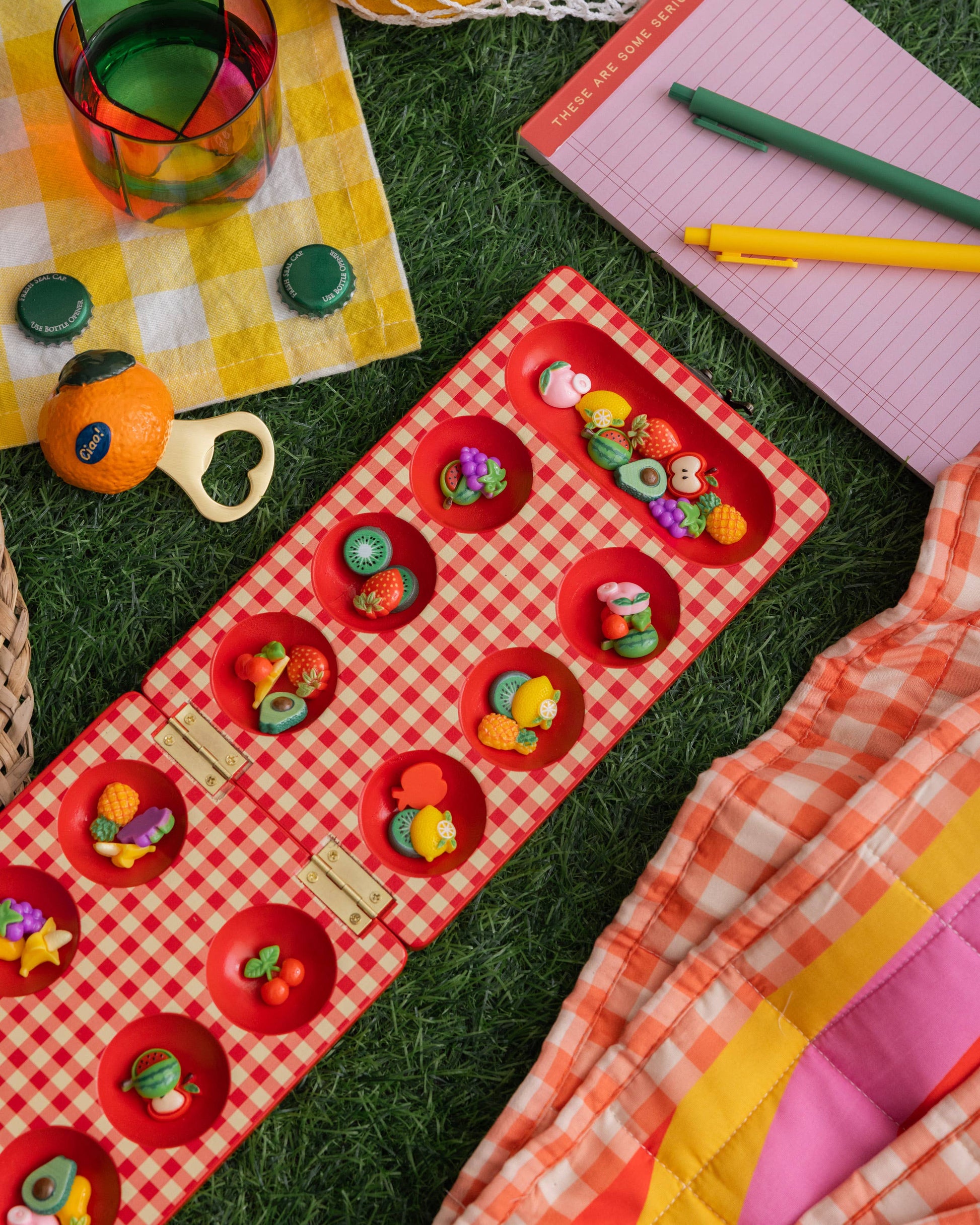 Colorful picnic setup with a red checkered blanket, toys, and stationery on grass.