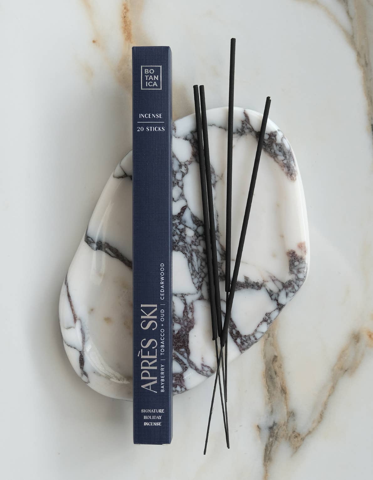 A dark navy rectangular box of Incense by Ritualica stands upright on a white marble tray with black veining, with several thin black incense sticks resting on top, all on a light marble surface.