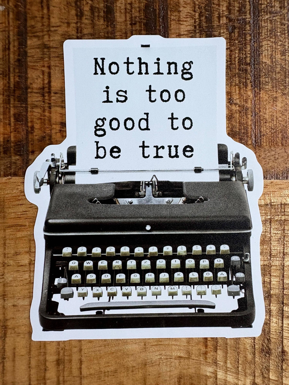 A die-cut sticker shaped like a vintage typewriter with a white sheet of paper that reads “Nothing is too good to be true” in black typewriter font, placed on a brown wood surface.