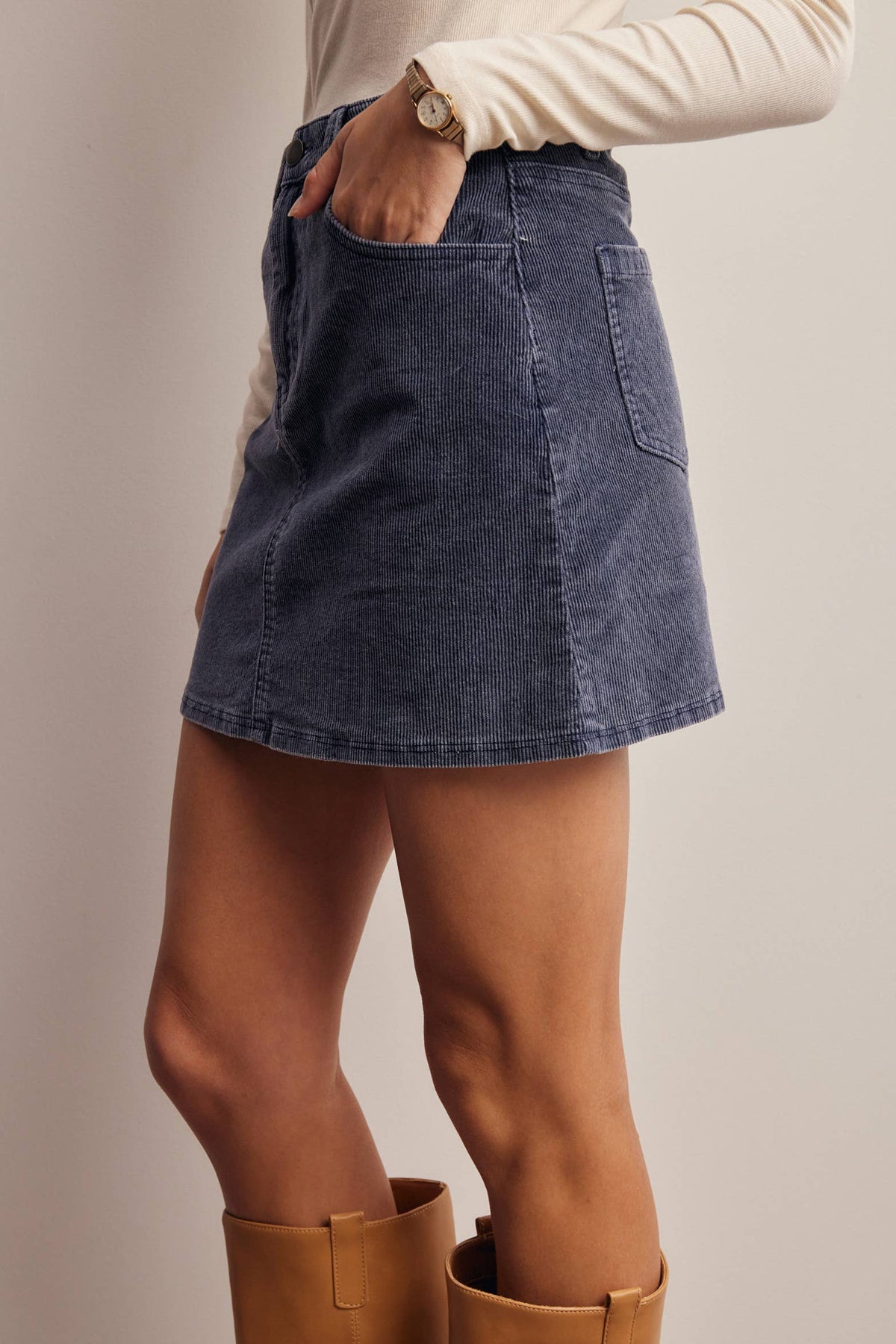 A person wearing a dark blue denim mini skirt with side pockets, paired with a cream long-sleeve top and tan boots, shown from the waist down against a plain beige studio background.