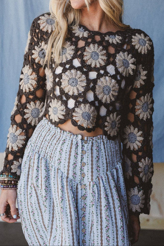 A woman wearing a black crochet long-sleeve crop top with beige floral patterns and a light blue patterned skirt, standing against a blue fabric backdrop.