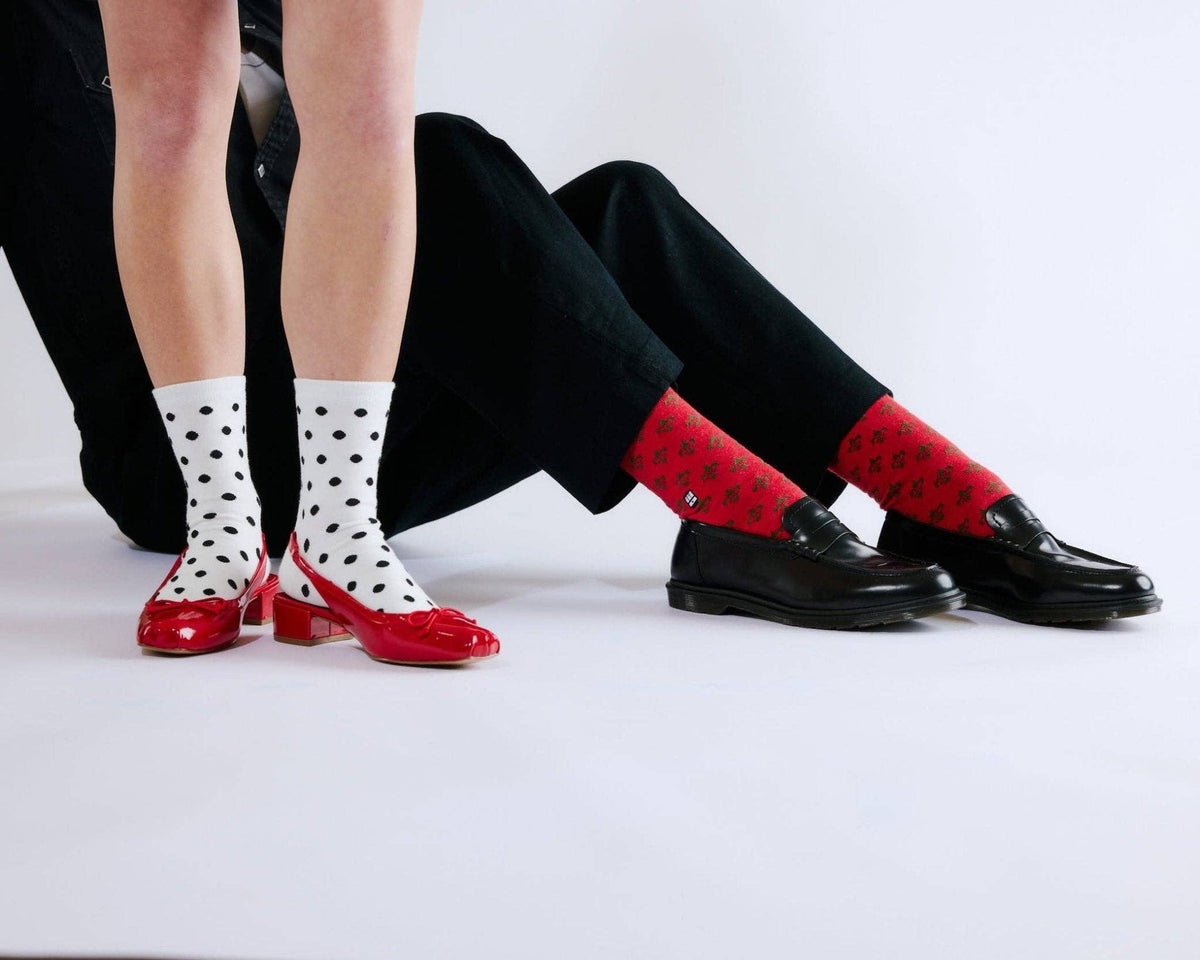 Three pairs of socks and shoes shown on models' lower legs and feet against a plain light background. One pair is white with black polka dots and red heels, one pair is red with a small white monogram, and one pair is solid black with black loafers.