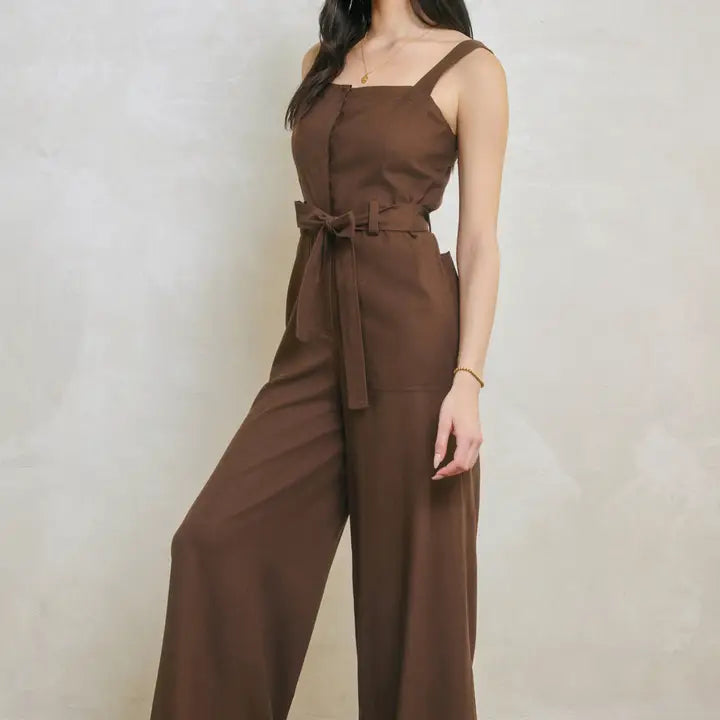 A woman wearing a brown sleeveless jumpsuit with wide legs, a square neckline, and a matching fabric tie belt at the waist, posed against a textured light beige wall.