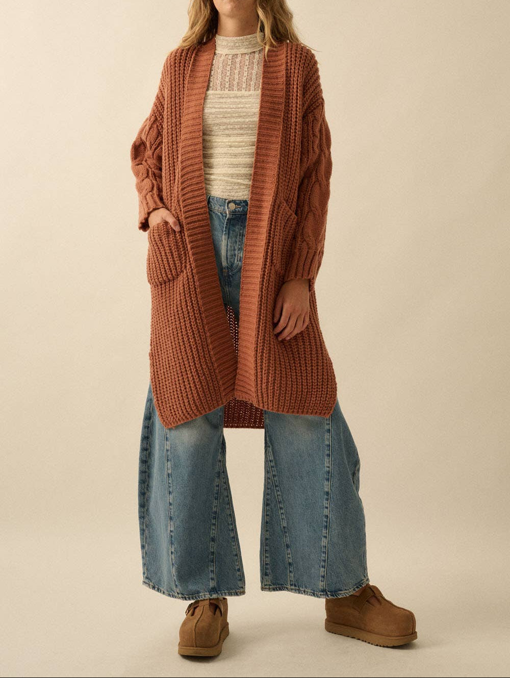 A woman modeling a long, rust-orange cable-knit cardigan over a cream ribbed top, blue wide-leg jeans, and brown boots, set against a plain beige studio background.