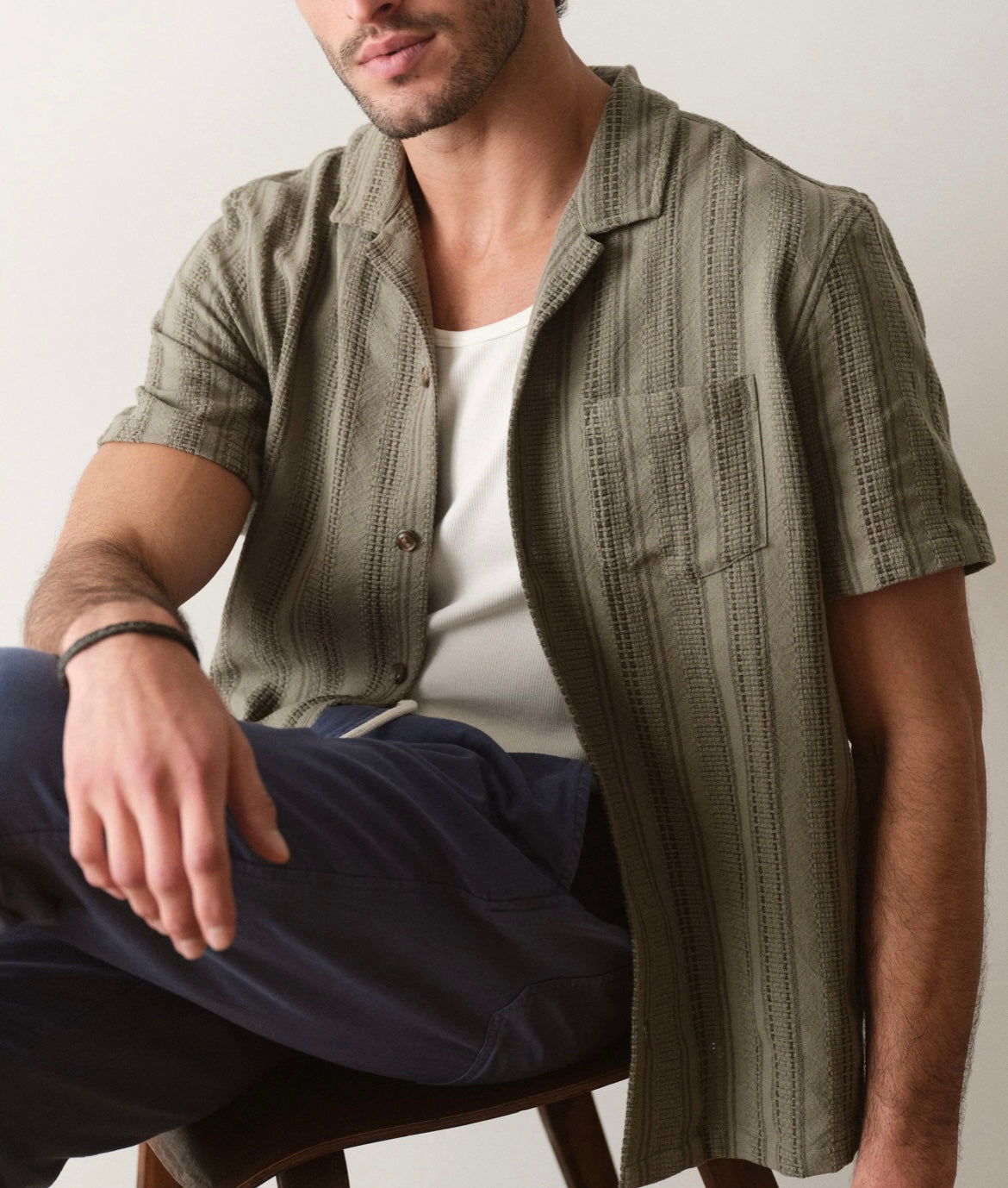 Man wearing a patterned green shirt over a white t-shirt, sitting on a chair.