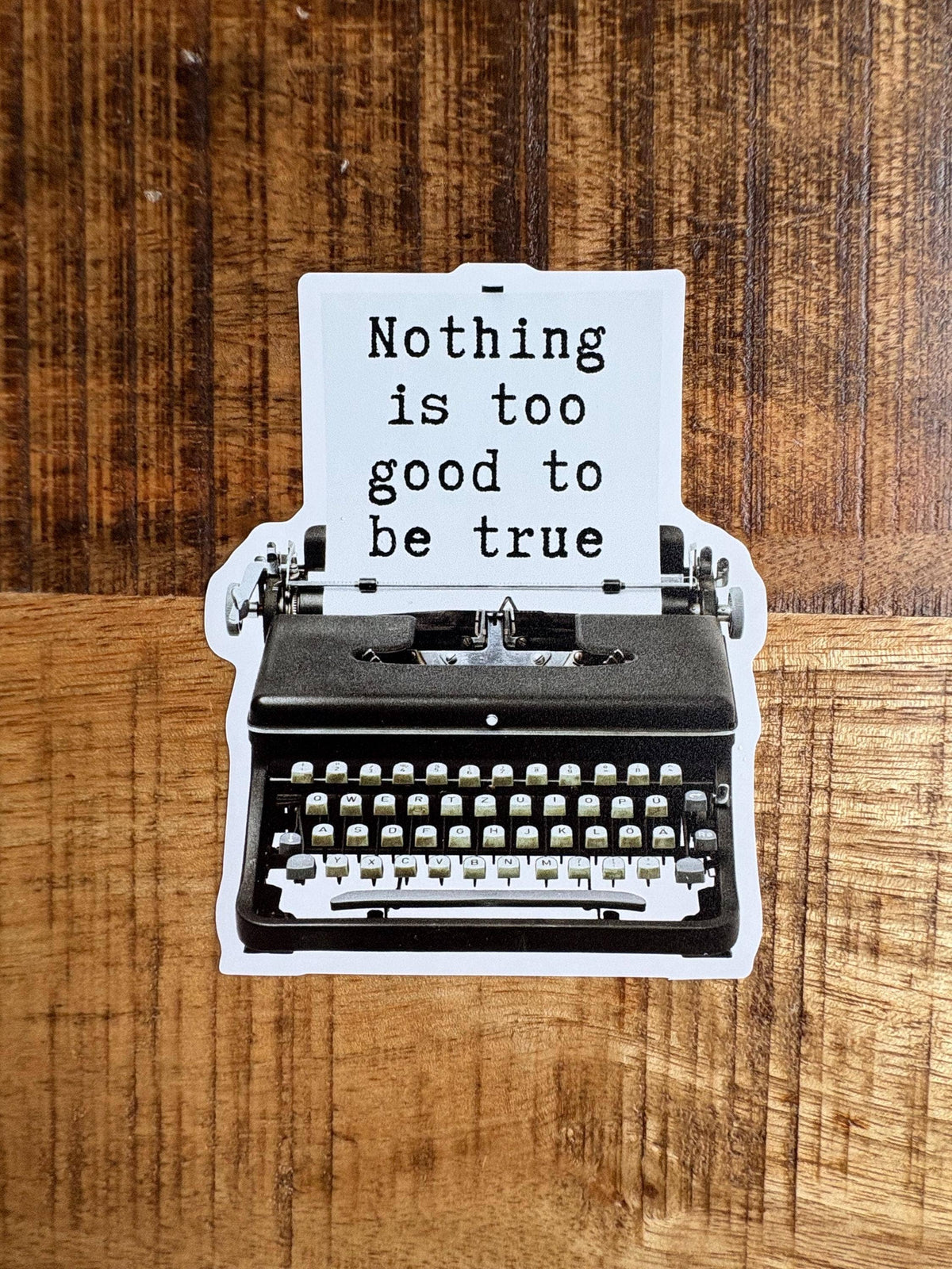 A die-cut sticker shaped like a vintage typewriter with a white sheet of paper that reads 'Nothing is too good to be true' in black typewriter font, placed on a wooden surface.