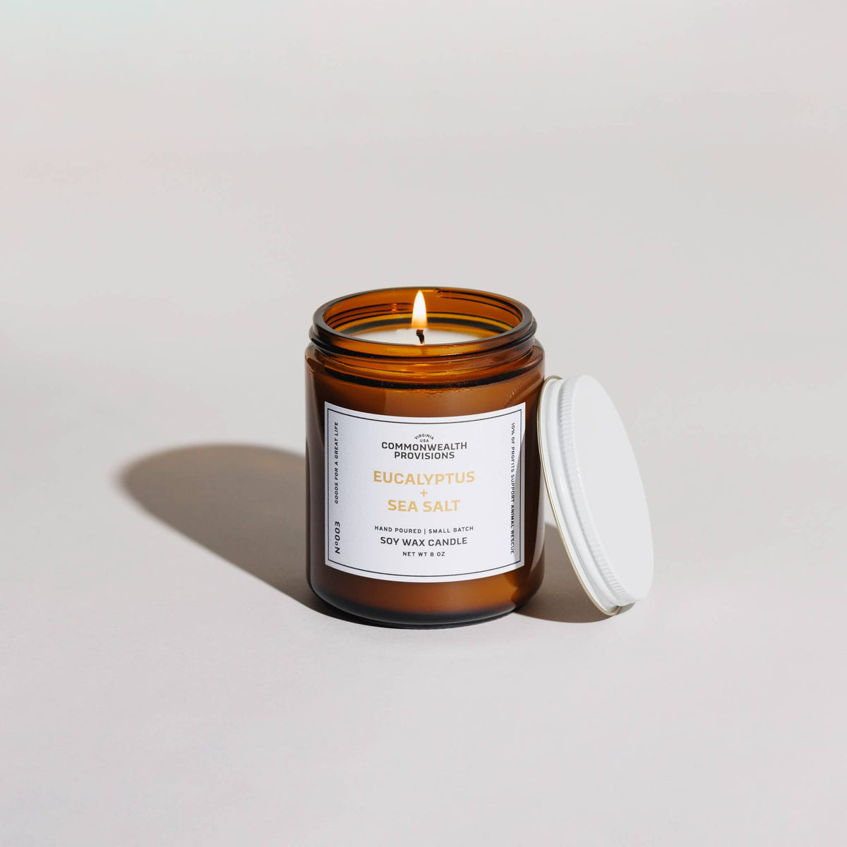 An amber glass jar candle with a white label reading 'Eucalyptus + Sea Salt' and a lit flame, shown on a light neutral background with the white lid resting beside the jar.