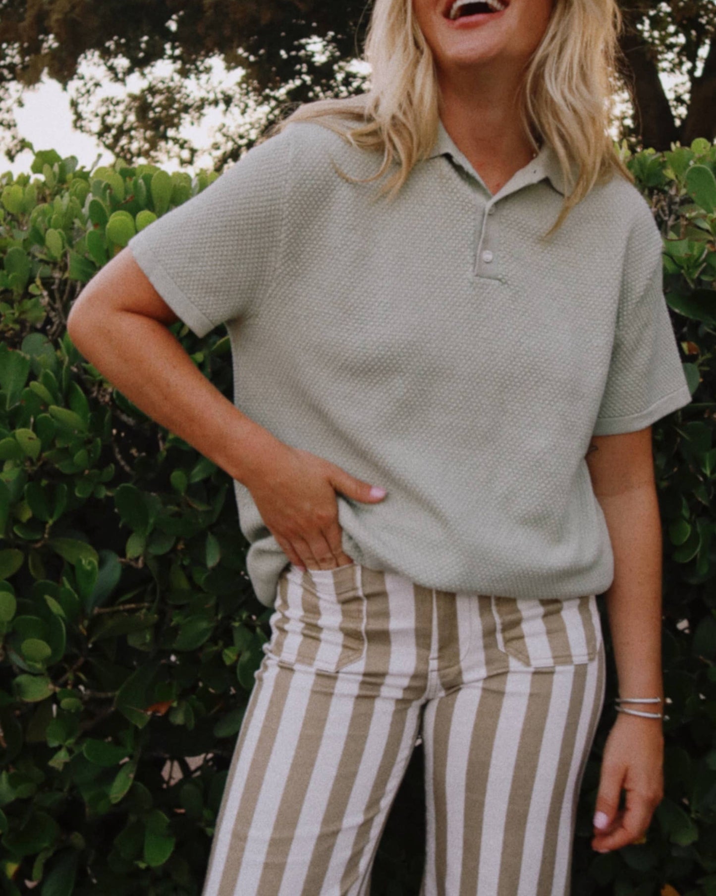Woman in a light green polo shirt and striped pants standing outdoors with greenery in the background